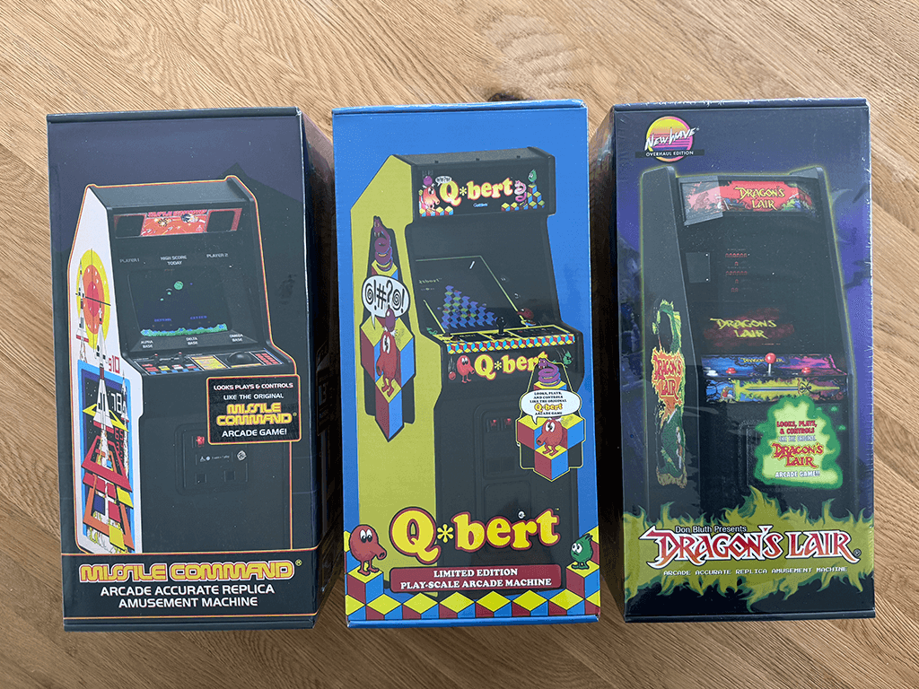 Three boxed New Wave 1/6 scaled arcade cabinets, Missle Command, Qbert and Dragons Liar on a wooden table top surface.