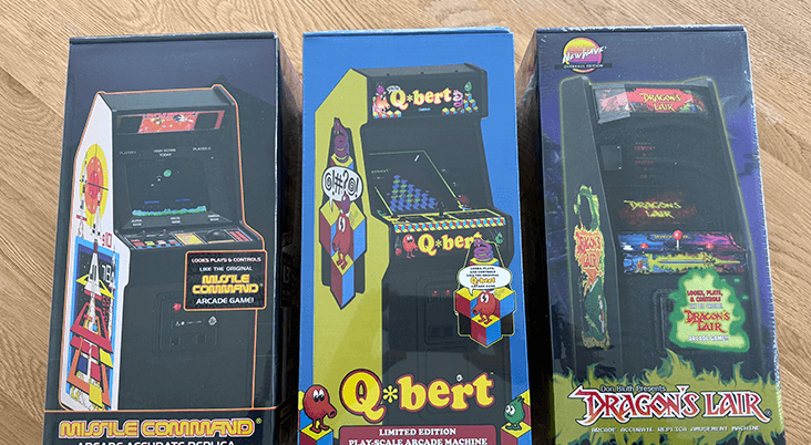 Three boxed New Wave 1/6 scaled arcade cabinets on a wooden table top surface.